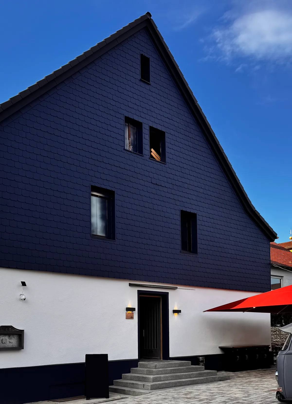 Der Landgasthof Engel in Hohenwart von der Straße aus: Giebel mit Schieferfassade, weißes Erdgeschoss mit Eingangstreppe und ein roter Sonnenschirm