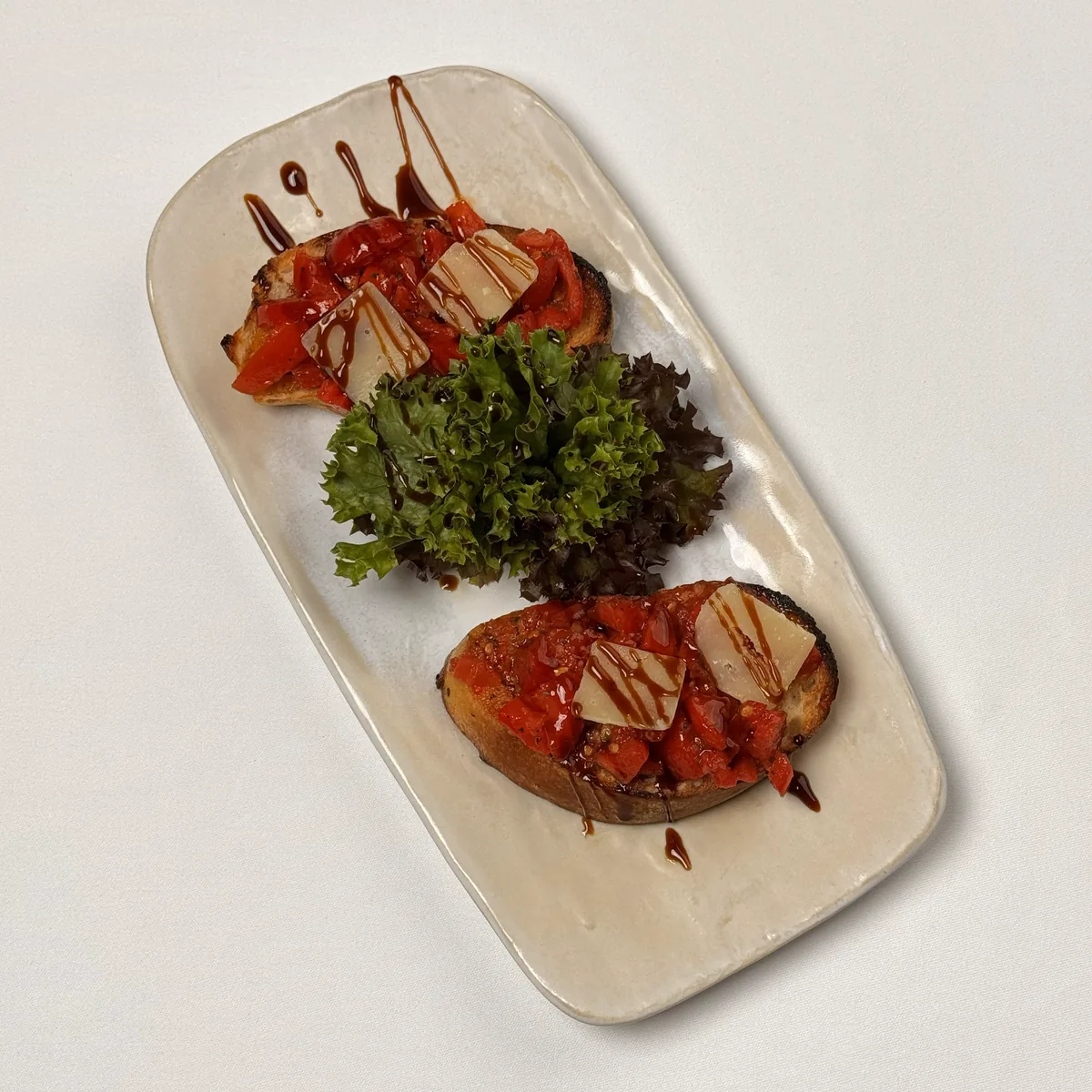 Zwei Bruschette mit Tomatenwürfeln, Parmesanspänen und Balsamico, dazwischen Blattsalat, auf einer länglichen Keramikplatte