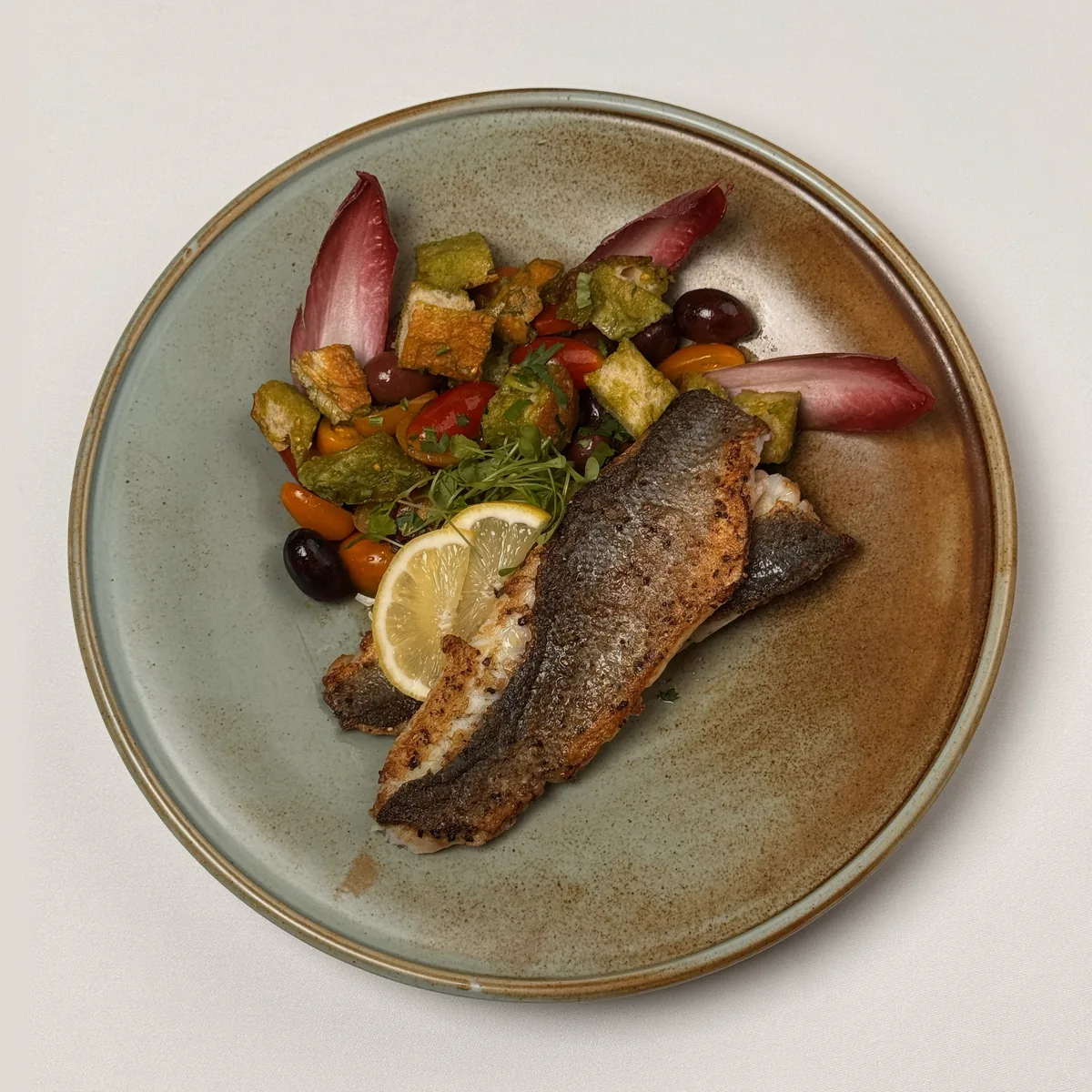 Zwei gebratene Wolfsbarsch-Filets auf der Haut mit Zitronenscheiben, Brotwürfeln, bunten Kirschtomaten, Kalamata-Oliven und rotem Chicorée